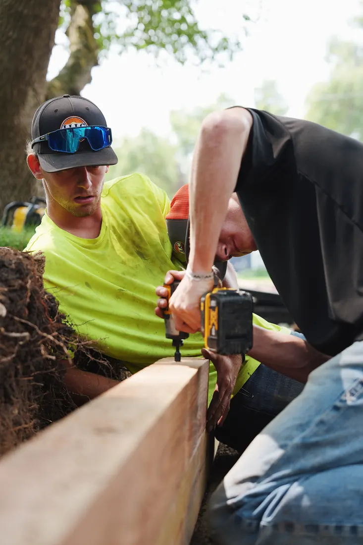 Two crew members drilling and pinning a timber retaining wall